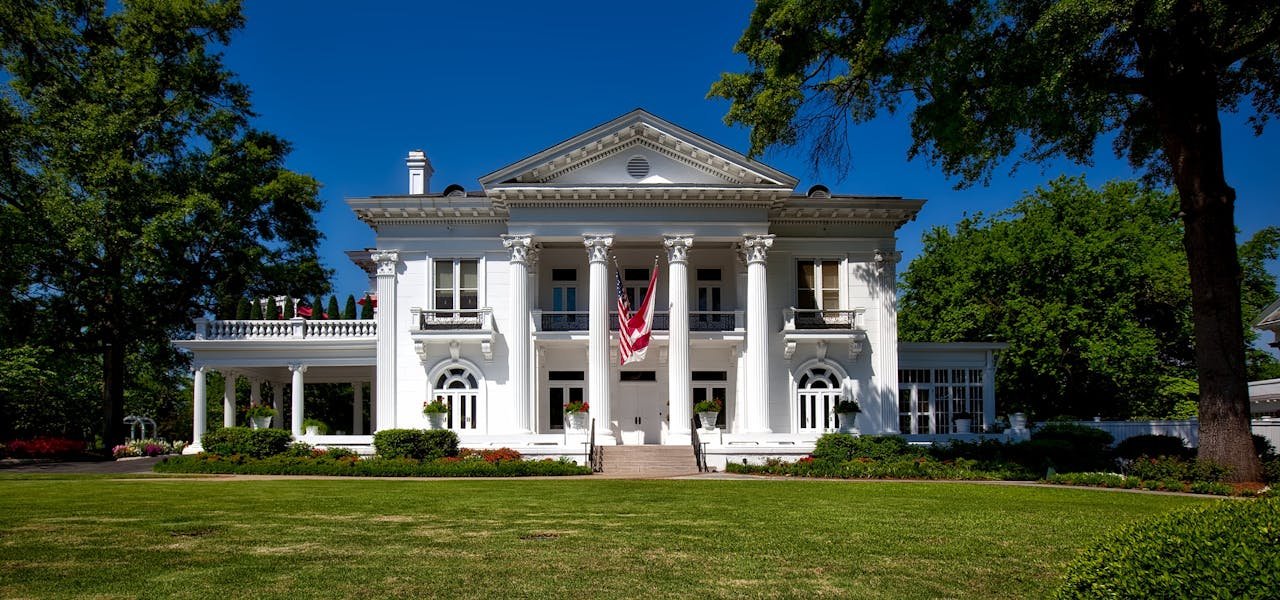A grand neoclassical mansion with lush gardens and American flag in a sunny setting.